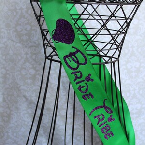 Puede incluir: Sash de sat&eacute;n verde con texto en purpurina morada que dice "Bride Tribe" y un coraz&oacute;n de purpurina morada.