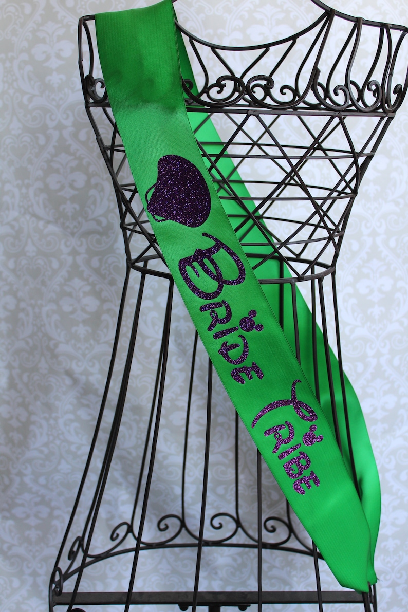 Puede incluir: Sash de sat&eacute;n verde con texto en purpurina morada que dice "Bride Tribe" y un coraz&oacute;n de purpurina morada.