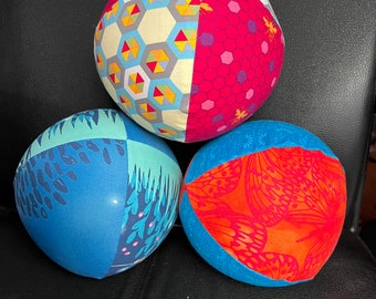Peut inclure: Trois boules rondes recouvertes de tissu avec des motifs différents. La boule du haut présente un motif géométrique jaune, bleu et rouge. La boule de gauche présente un motif bleu et vert avec des accents blancs. La boule de droite présente un motif bleu et orange avec des accents blancs.