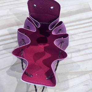 May include: A deep magenta drawstring bag with a contrasting red interior. The bag has a unique, scalloped edge design and is open, revealing the interior. The drawstring is a dark brown colour.