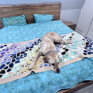 May include: A beige dog lies on a bed with a blue patterned bedspread and matching cushions. The bed has a wooden headboard and a decorative runner with blue, purple, and black stone-like shapes.