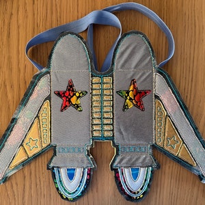 Puede incluir: Un disfraz de nave espacial de fieltro gris con tirantes azules, ribetes dorados y azules y estrellas de colores.