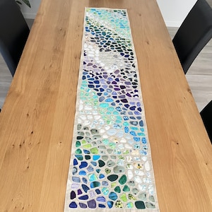 Może przedstawiać: Bieżnik stołowy z mozaikowym wzorem z niebieskich, zielonych i fioletowych kawałków tkaniny. Bieżnik leży na drewnianym stole.