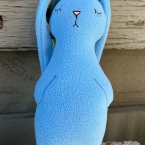 May include: A blue plush bunny with long ears, closed eyes, and a pink nose. The bunny is sitting on a wooden surface.