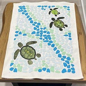 May include: White rectangular decorative towel featuring three sea turtles in shades of green and black. The background is a mosaic of blue, light blue, and green shapes, resembling sea glass. The towel is placed on a wooden table.