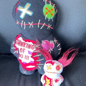 May include: Two voodoo dolls, one black with red and green accents and the words "Thinking of You!!!" and the other white with red accents. The dolls are stitched together with red thread and have a red heart on the front.