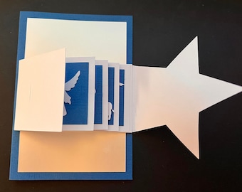 Peut inclure: Une carte pop-up bleue et blanche avec un rabat en forme d'étoile. La carte présente une série de silhouettes blanches d'anges et d'autres figures religieuses.