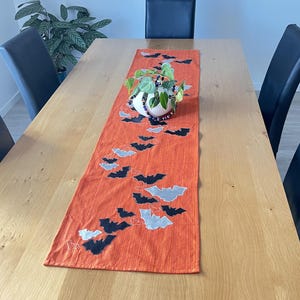 Puede incluir: Un camino de mesa naranja con un diseño de murciélagos negros y grises. El camino de mesa está colocado sobre una mesa de madera clara. Un jarrón blanco con una planta se encuentra sobre el camino de mesa. Sillas negras rodean la mesa.