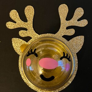 Peut inclure: Une d&eacute;coration de renne en paillettes dor&eacute;es avec un d&ocirc;me en plastique transparent et un visage souriant rose et noir.