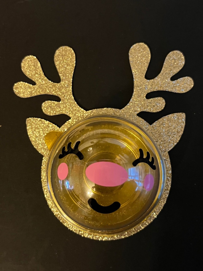 Peut inclure: Une d&eacute;coration de renne en paillettes dor&eacute;es avec un d&ocirc;me en plastique transparent et un visage souriant rose et noir.