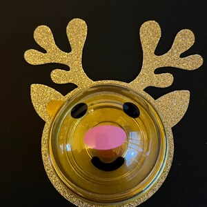 Peut inclure: Une d&eacute;coration de renne en paillettes dor&eacute;es avec un d&ocirc;me en plastique transparent contenant un visage de renne jaune et noir avec un nez rose.