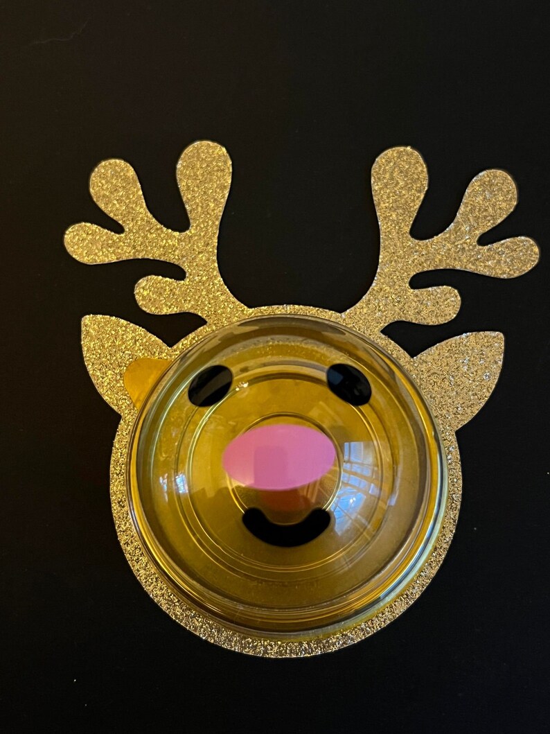 Peut inclure: Une d&eacute;coration de renne en paillettes dor&eacute;es avec un d&ocirc;me en plastique transparent contenant un visage de renne jaune et noir avec un nez rose.