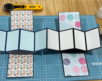Peut inclure: Deux cartes ouvertes de style accordéon avec du papier à motifs sur le devant et le dos. La carte de gauche a un fond bleu avec une bordure blanche et la carte de droite a un fond noir avec une bordure blanche. La carte de gauche a un motif de petits cercles colorés et la carte de droite a un motif de grands cercles colorés avec des lignes noires.