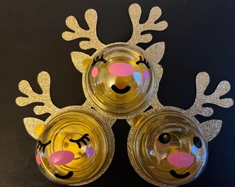 Peut inclure: Trois décorations de rennes dorées avec des accents de paillettes. Chaque décoration a un dôme en plastique transparent avec un visage de renne de dessin animé mignon avec des joues roses et des yeux noirs. 