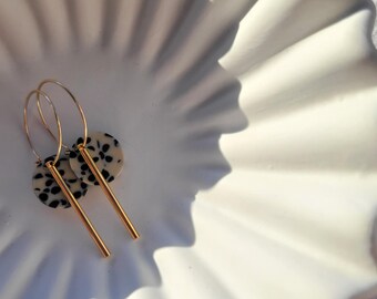 May include: Gold hoop earrings with a cream and black speckled disc and a gold bar pendant. The earrings are displayed on a white, ruffled, decorative plate.
