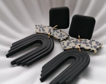 May include: A pair of black and white statement earrings with a geometric design. The earrings feature a black arch shape with a white and black patterned square on top.