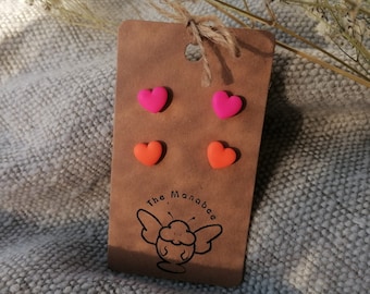 May include: A brown card with four heart-shaped earrings in bright pink and orange. The card has a string for hanging and a logo that says "The Manabee". The earrings are on a gray textured surface.