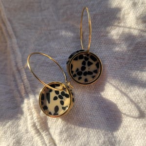 May include: A pair of gold hoop earrings with circular pendants. The pendants feature a cream-colored background with black speckled patterns, encased in a gold frame. The earrings are displayed on a white, textured surface.