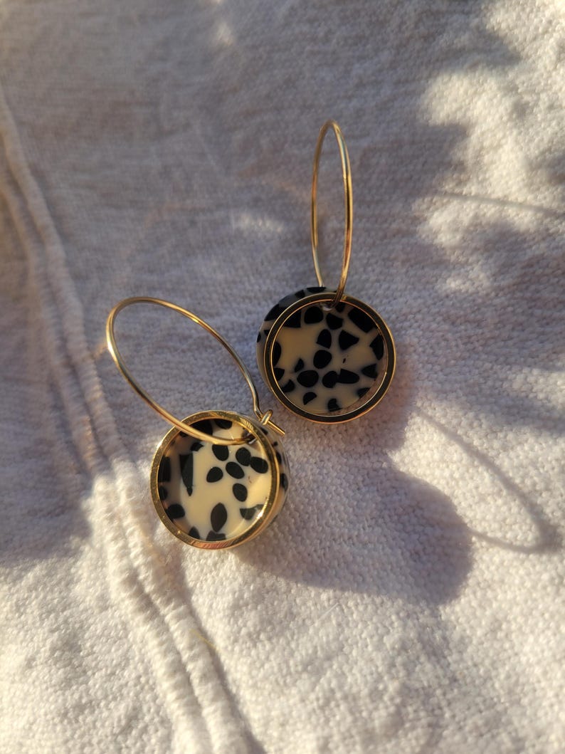 May include: A pair of gold hoop earrings with circular pendants. The pendants feature a cream-colored background with black speckled patterns, encased in a gold frame. The earrings are displayed on a white, textured surface.