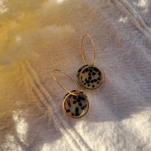 May include: A pair of gold hoop earrings with circular pendants. The pendants feature a speckled black and white pattern, set within a gold frame. The earrings are displayed on a white, textured surface.