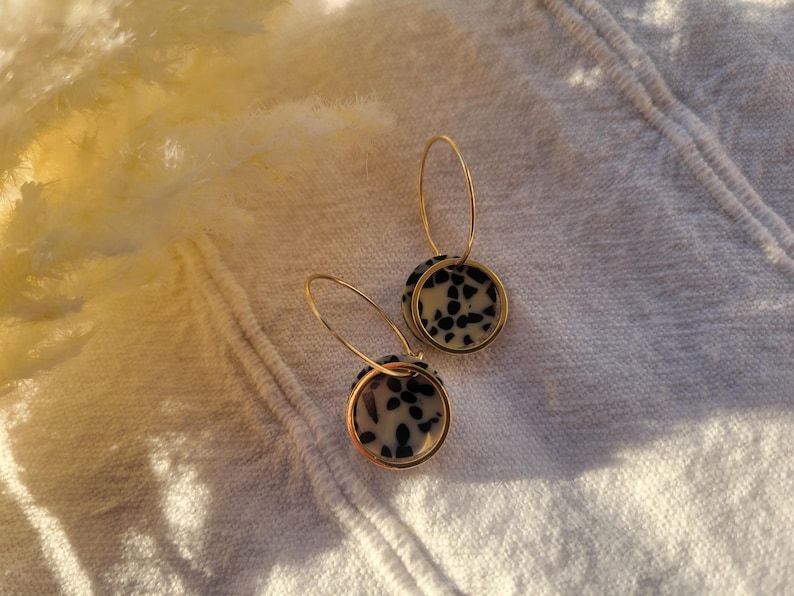 May include: A pair of gold hoop earrings with circular pendants. The pendants feature a speckled black and white pattern, set within a gold frame. The earrings are displayed on a white, textured surface.