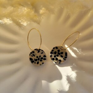 May include: A pair of gold hoop earrings with circular, cream-colored pendants featuring black spots. The earrings are displayed on a white surface, with soft, feathery elements in the background.
