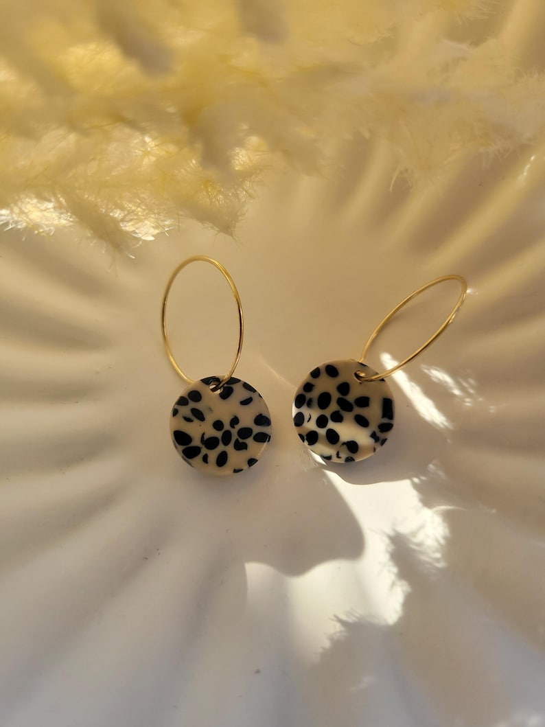 May include: A pair of gold hoop earrings with circular, cream-colored pendants featuring black spots. The earrings are displayed on a white surface, with soft, feathery elements in the background.