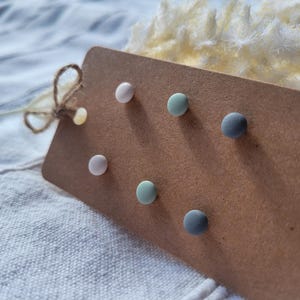Könnte beinhalten: Ein Set aus sechs Ohrsteckern in Pastellfarben: Rosa, Mintgrün und Grau. Die Ohrringe sind auf einem braunen Pappanhänger mit einer Schnur-Schleife präsentiert. Der Hintergrund zeigt weißen Stoff und einen flauschigen, cremefarbenen Akzent.