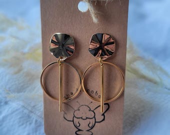 May include: A pair of gold-toned dangle earrings featuring a textured, scalloped circular top piece connected to a smooth, thin vertical bar centered within a larger, plain gold-toned circular hoop. The earrings are displayed on a brown card.