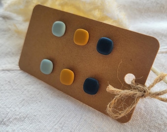 May include: A set of six small, square stud earrings with rounded corners, arranged in two rows on a brown rectangular display card. The earrings feature a matte finish in three distinct colors: light blue, mustard yellow, and dark navy blue.