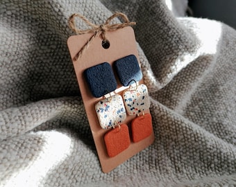 May include: A pair of square dangle earrings in navy blue, speckled white, and burnt orange. The earrings are attached to a brown tag with a twine bow. The earrings are made of polymer clay and have gold-colored earring hooks.
