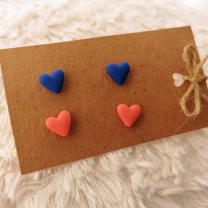 Könnte beinhalten: Vier herzförmige Ohrringe auf einer braunen Karte. Zwei Ohrringe sind blau und zwei sind korallenfarben.