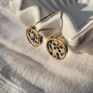 May include: A pair of gold-toned earrings with a circular design featuring a black and cream spotted pattern. The earrings are displayed on a white, shell-shaped dish, creating a contrast of textures and colors.