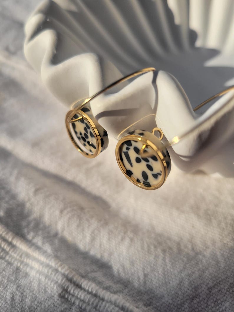 May include: A pair of gold-toned earrings with a circular design featuring a black and cream spotted pattern. The earrings are displayed on a white, shell-shaped dish, creating a contrast of textures and colors.