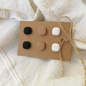 Könnte beinhalten: Ein Set aus sechs quadratischen Ohrsteckern in Schwarz, Beige und Weiß, präsentiert auf einer braunen Karte, die mit Bindfaden gebunden ist. Die Ohrringe sind in zwei Dreierreihen angeordnet und zeigen ein minimalistisches Design.