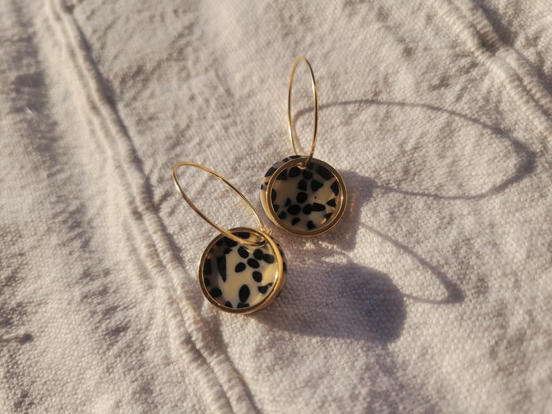 May include: A pair of gold hoop earrings with circular pendants. The pendants feature a cream-colored background with black speckled patterns, framed by a gold border. The earrings are displayed on a white, textured surface.