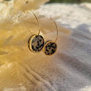 May include: Gold-tone hoop earrings with circular pendants. The pendants feature a dalmatian-like pattern of black spots on a cream background, framed by a gold border. The earrings are displayed against a soft, textured background.