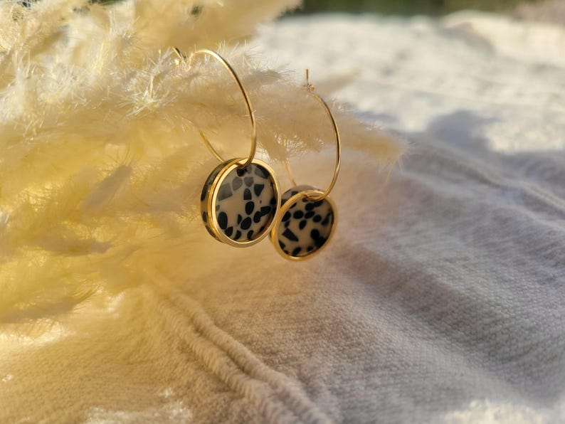May include: Gold-tone hoop earrings with circular pendants. The pendants feature a dalmatian-like pattern of black spots on a cream background, framed by a gold border. The earrings are displayed against a soft, textured background.