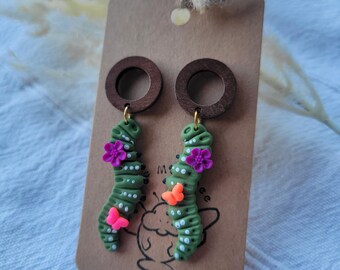May include: A pair of dangle earrings featuring green caterpillar-shaped charms made of clay. Each caterpillar is decorated with small white dots, a bright pink flower, and a tiny orange butterfly. The charms hang from dark brown wooden rings.