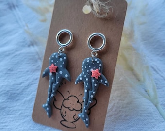 May include: A pair of dangle earrings featuring grey whale sharks with white polka dots and a small coral-colored starfish attached to each shark. The earrings hang from silver-toned circular hoops and are displayed on a brown card.