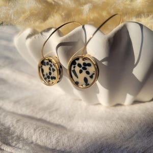 May include: A pair of gold-tone hoop earrings with circular pendants. The pendants feature a cream-colored background with black speckled patterns, framed by a gold border. The earrings are displayed in a white, scalloped bowl.