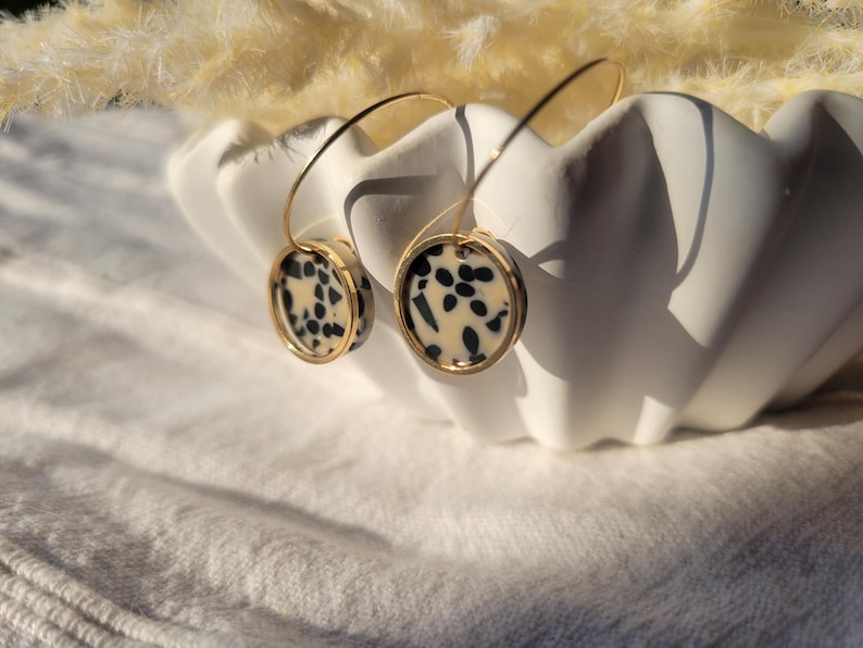 May include: A pair of gold-tone hoop earrings with circular pendants. The pendants feature a cream-colored background with black speckled patterns, framed by a gold border. The earrings are displayed in a white, scalloped bowl.
