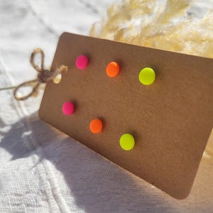 Könnte beinhalten: Ein braunes rechteckiges Etikett mit sechs bunten, runden Reißzwecken in Neonrosa, Orange und Gelb. Das Etikett ist mit einer natürlichen Schnur-Schleife gebunden. Der Hintergrund ist eine weiße, strukturierte Oberfläche.