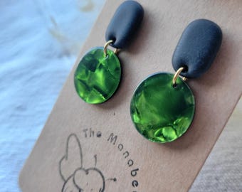 May include: A pair of dangle earrings featuring matte black oval studs connected by gold-toned jump rings to circular, translucent green acrylic charms with a marbled texture. The earrings are displayed on a brown card labeled The Manabee.
