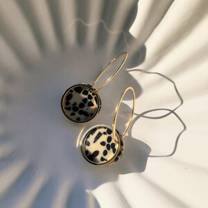 May include: A pair of gold hoop earrings with circular pendants. The pendants feature a cream-colored background with black spots, framed by a gold border. The earrings are set against a white, textured surface.