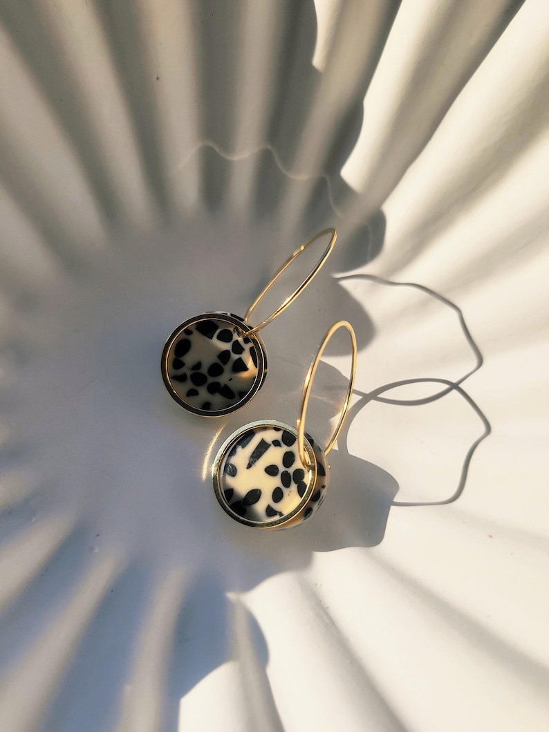 May include: A pair of gold hoop earrings with circular pendants. The pendants feature a cream-colored background with black spots, framed by a gold border. The earrings are set against a white, textured surface.