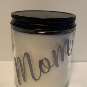 May include: A clear glass candle jar with a black lid. The jar contains white wax and has the word "Mom" in gray script on the front. The candle is a simple, elegant design.