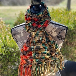 May include: A handmade crochet scarf in shades of brown, orange, and green, displayed on a mannequin. The scarf features a fringed edge and a textured pattern. The color palette includes warm tones, creating a cozy and stylish accessory.