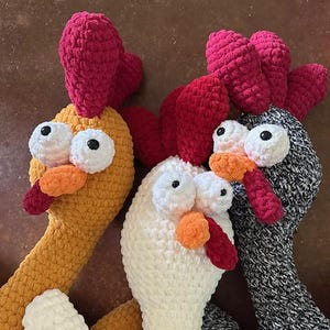 Puede incluir: Tres juguetes de gallo de crochet. Uno es naranja con una cresta y una barbilla rojas, otro es blanco con una cresta y una barbilla rojas, y el último es gris con una cresta y una barbilla rojas.
