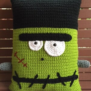May include: Green crocheted pillow with a Frankenstein face. The pillow has black eyes, a black mouth, and black stitches around the neck.